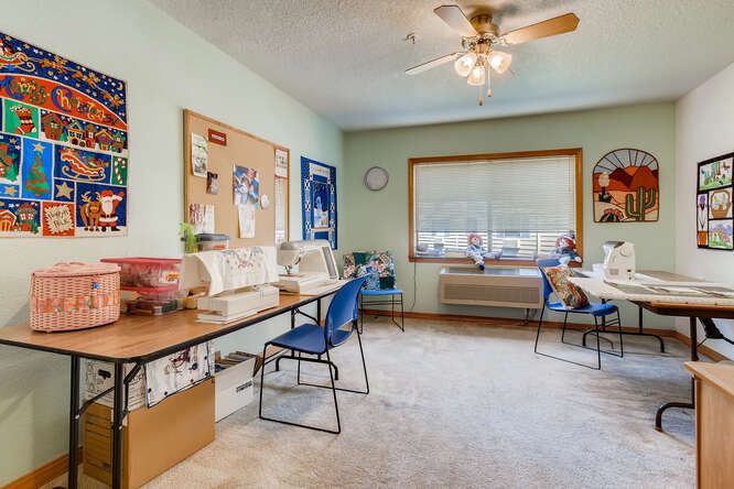 A well-organized craft room featuring sewing machines, a bulletin board, and ample workspace with chairs, illuminated by natural light from a large window.