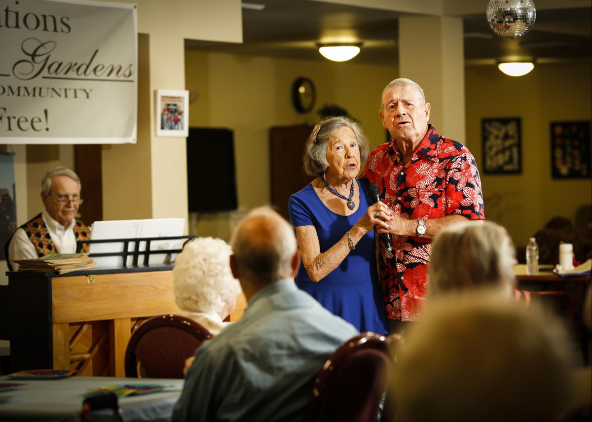 An elderly couple performs in front of an audience at an indoor event.