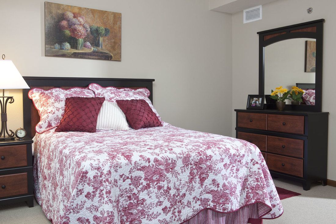 A cozy bedroom featuring a bed with floral bedding and decorative pillows, alongside a wooden dresser and mirror.