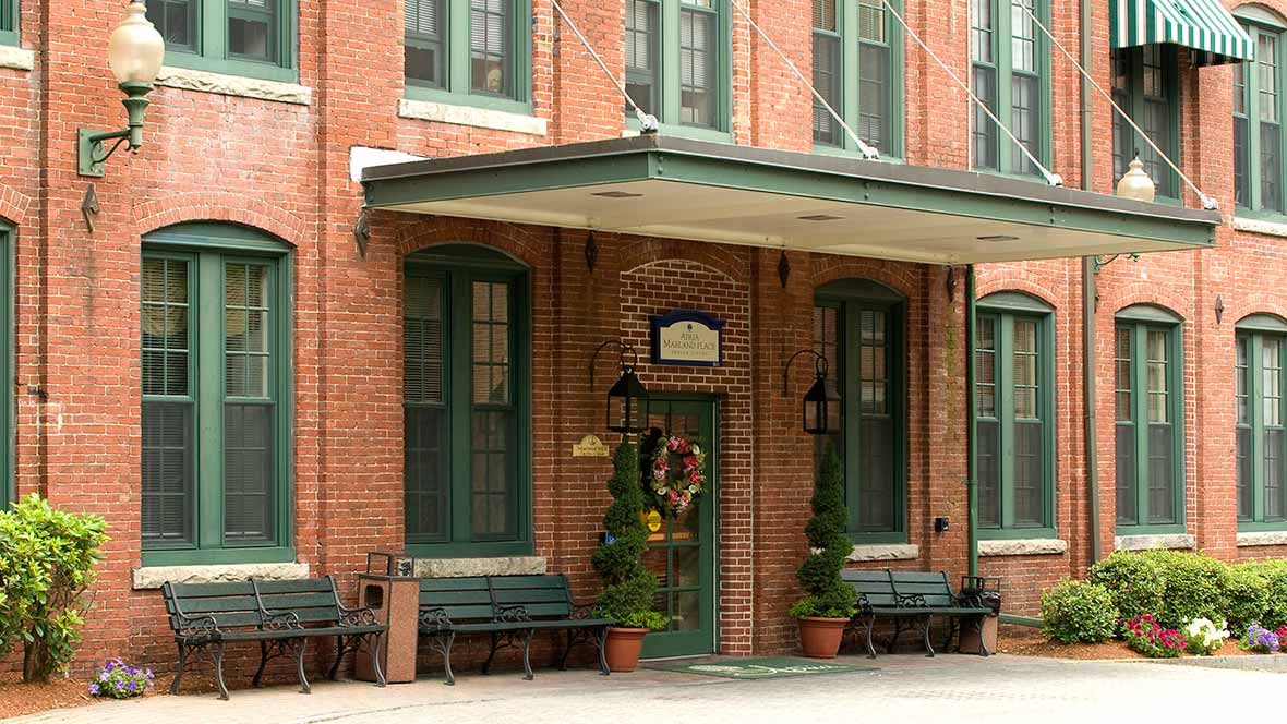 A charming historic brick building with green accents, featuring an entrance adorned with potted plants and a wreath.