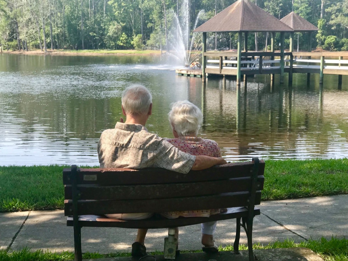 An elderly couple sitting on a bench by the lake, enjoying the scenic view of a park with a fountain.