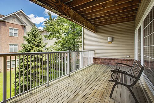 A spacious wooden balcony with outdoor seating, overlooking trees in a residential area.