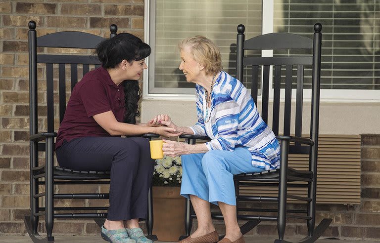 A caregiver and an elderly woman share a conversation while seated on rocking chairs outside.