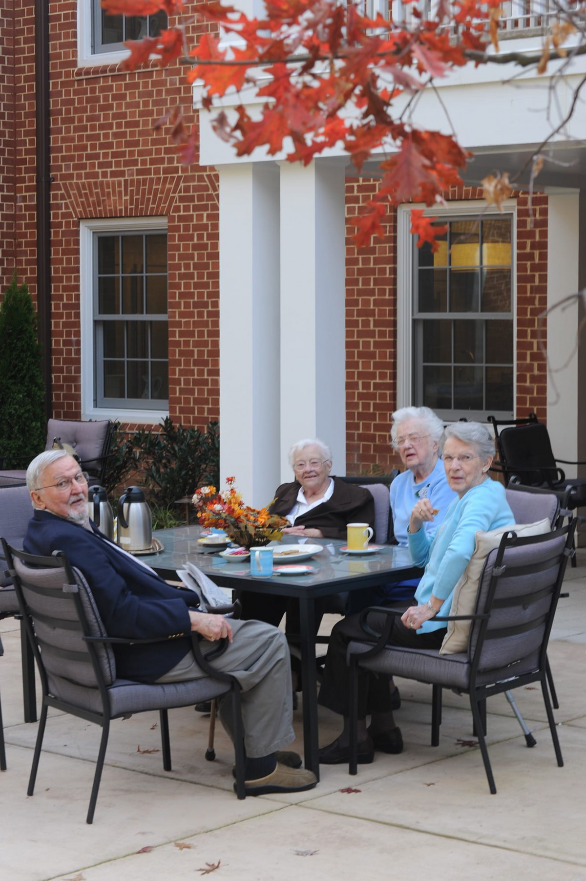 A group of seniors enjoying a social gathering outdoors during autumn, sitting at a table near a brick building.