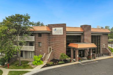 A two-story commercial building with a sign, surrounded by greenery and featuring a prominent entrance.