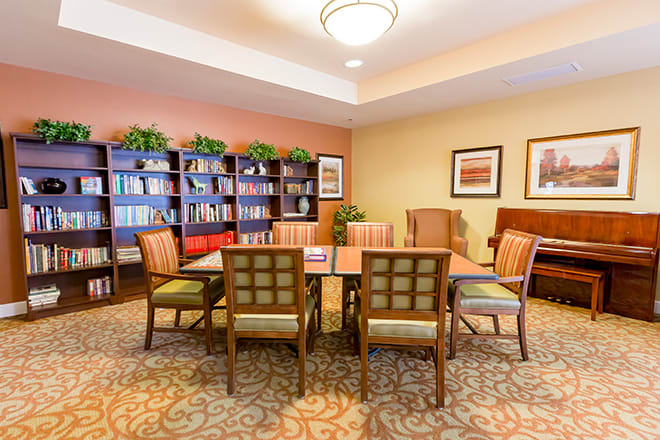 A well-lit library room with a central table and chairs, surrounded by bookshelves and decorative plants.