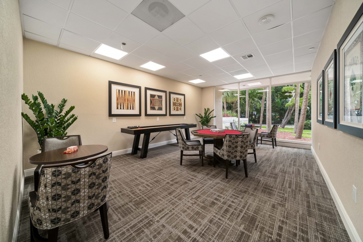 A modern game room with tables and chairs, decorated with artwork and plants, illuminated by natural light from large windows.