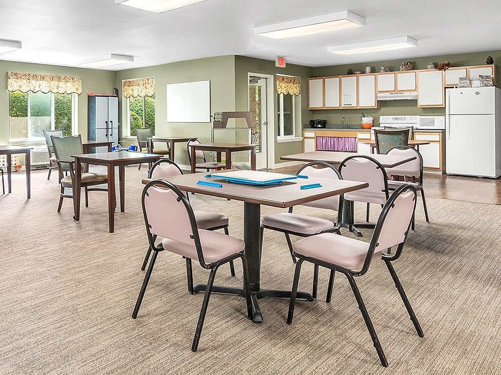 A communal dining area with tables and chairs, featuring a kitchen space in the background.