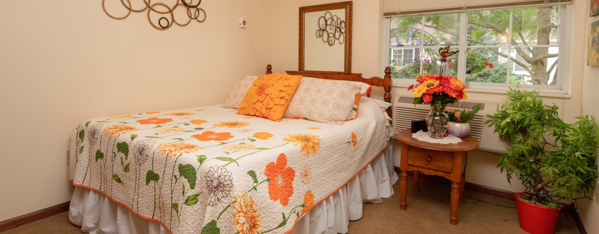 A cozy bedroom featuring a bed with a floral quilt and pillows, alongside a nightstand and mirror.