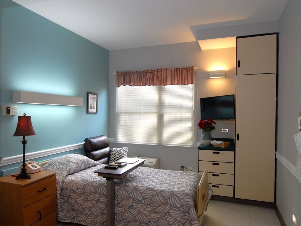 A neatly arranged hospital room with a single bed, TV, and furniture beneath the window.