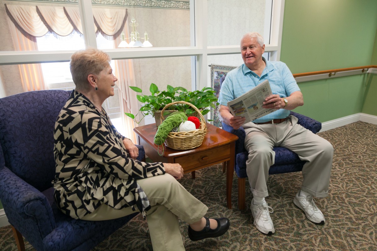 Two seniors enjoying a conversation indoors, with one reading a newspaper.