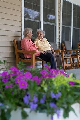 Two elderly women sitting on a porch having a conversation, with vibrant purple flowers in the foreground.