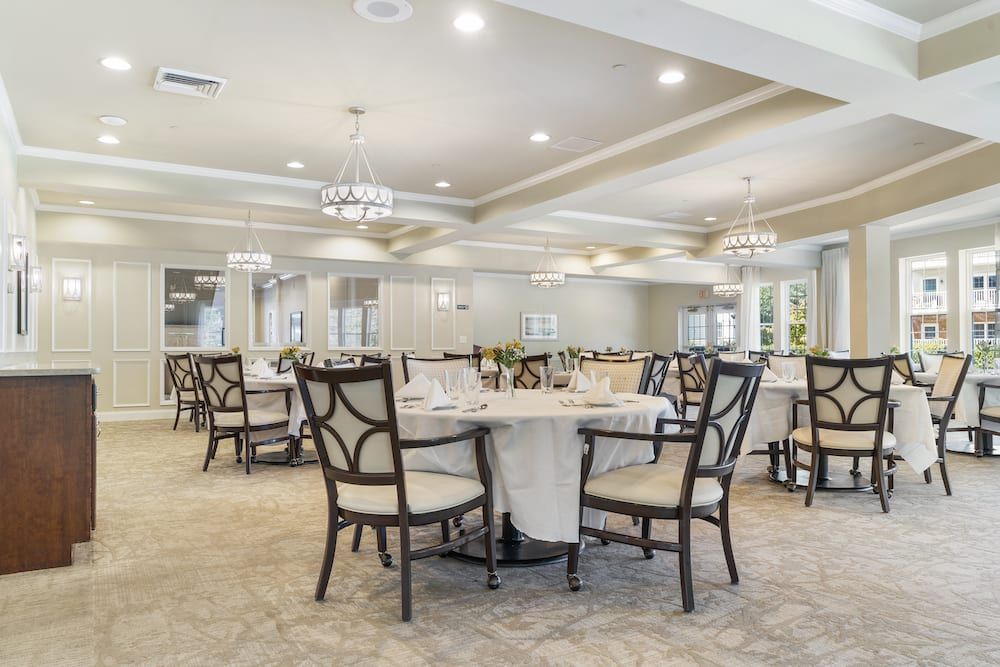 An elegantly designed dining room with round tables, upholstered chairs, and chandeliers, featuring bright lighting and a carpeted floor.