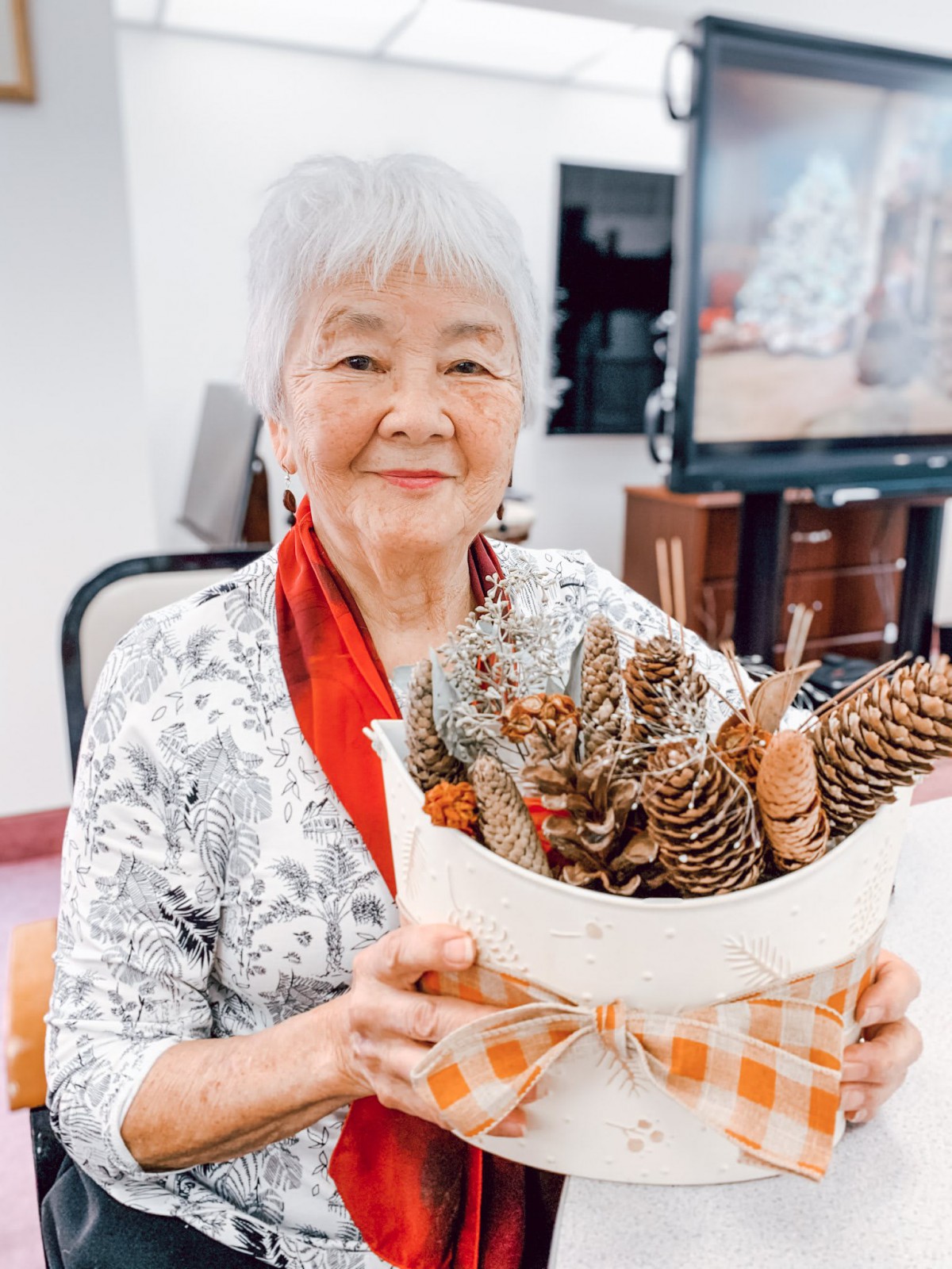 An elderly woman smiling while holding a decorative basket filled with pine cones.