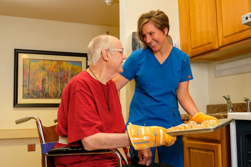 A caregiver assists an elderly person in a kitchen setting, both sharing a moment of joy.