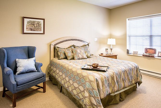 A cozy bedroom with a neatly made bed, an armchair, and a bedside table with a lamp.