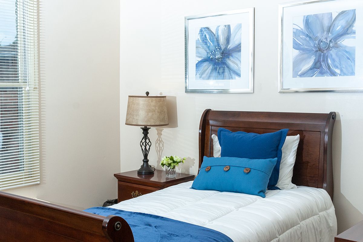 A neatly arranged bedroom with a single bed featuring blue decorative pillows and a runner, accompanied by a wooden nightstand with a lamp and floral artwork on the walls.