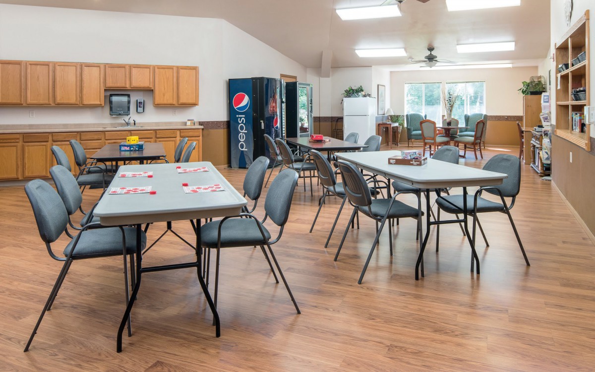 A spacious community center room with tables and chairs, a vending machine, and a small kitchen area.