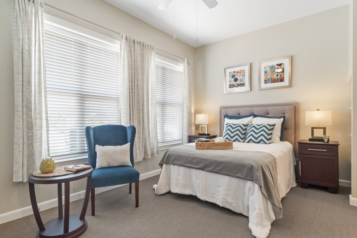 A cozy bedroom featuring a neatly made bed with decorative pillows, a blue armchair, and soft natural light streaming through large windows.