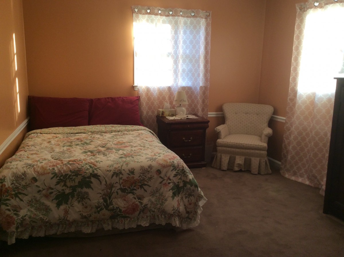 A cozy bedroom with a floral bedspread, two windows with curtains, a nightstand, and an upholstered chair.