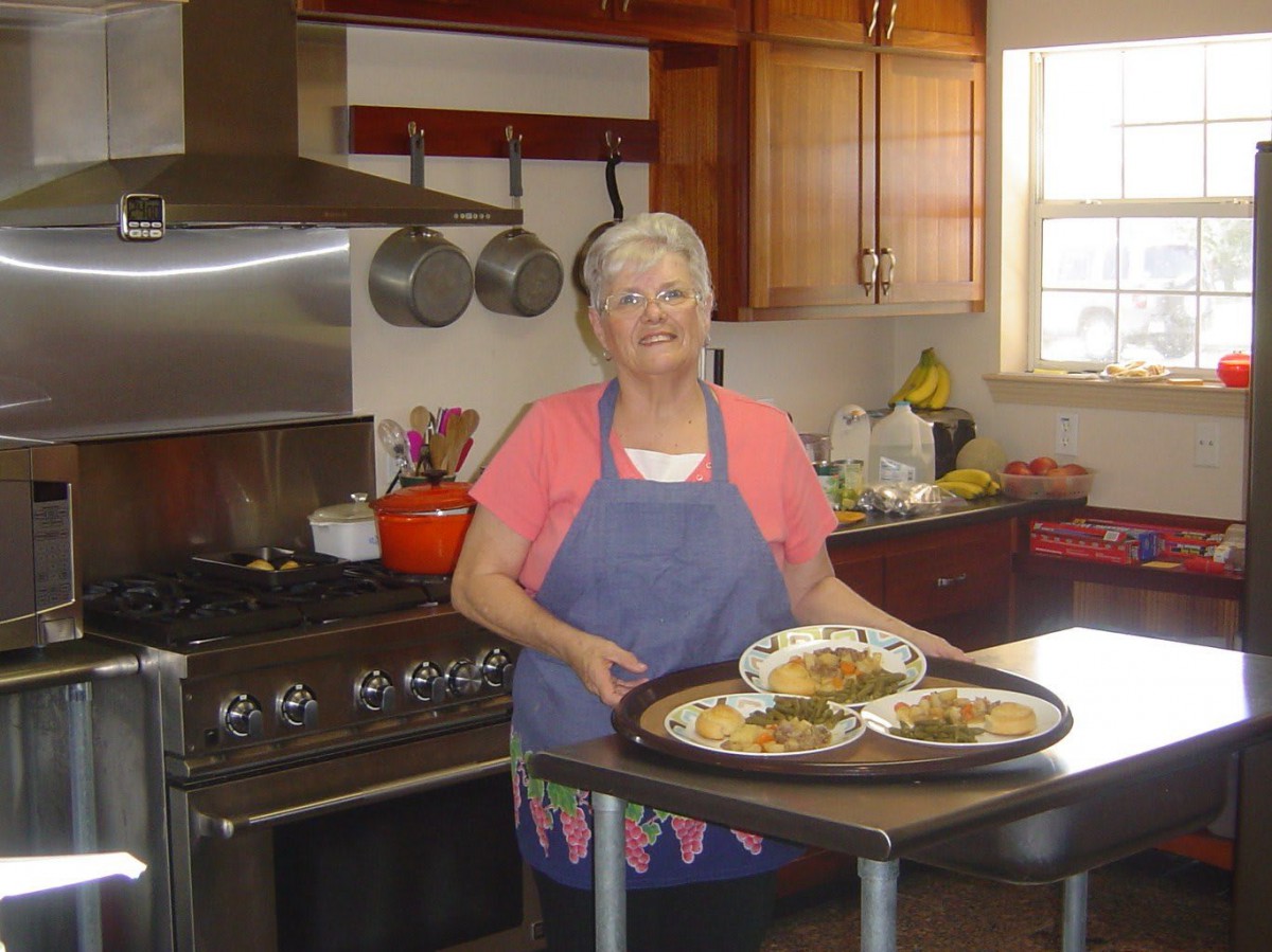 A smiling woman stands in a kitchen, holding a tray of food.