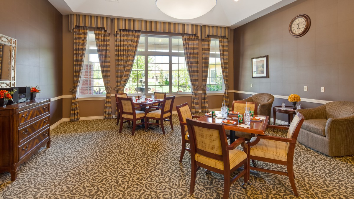 A cozy dining room with tables and chairs arranged on a patterned carpet, featuring large windows and elegant curtains.
