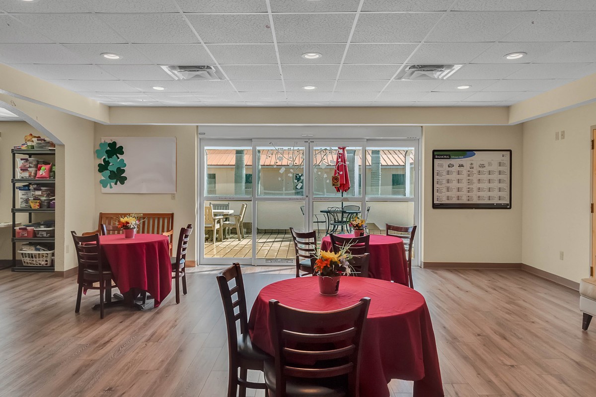 A cozy dining area with tables set with red tablecloths and chairs around them, featuring large windows for natural light.