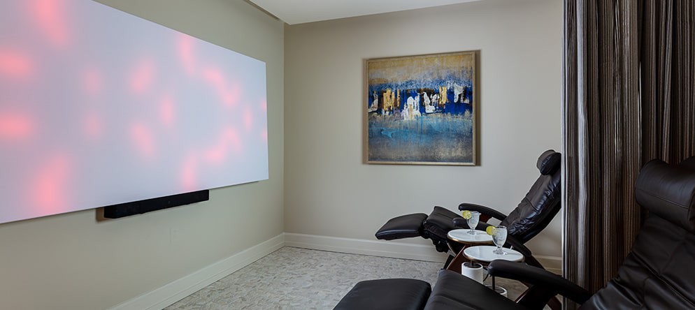 A modern home theater room featuring a projector screen and comfortable recliner chairs, adorned with an abstract cityscape painting on the wall.
