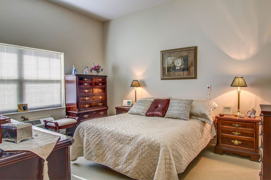 A cozy bedroom with a neatly made bed, adorned with pillows and a quilt, flanked by lamps on side tables. The room features elegant wooden furniture and wall art, creating a warm and inviting atmosphere.