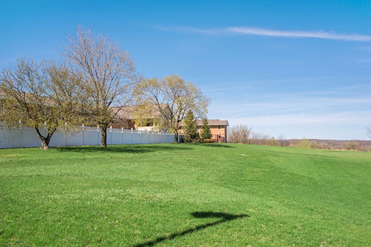 A sunny landscape featuring a house surrounded by trees and a fence, with green grass in the foreground.