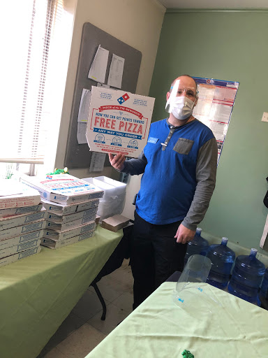 An individual wearing a mask holds a sign for free pizza at a community event, surrounded by boxes of pizza.
