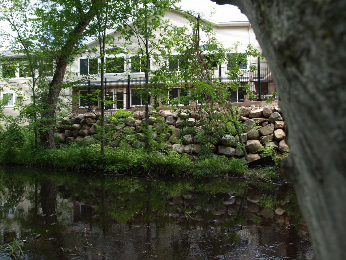 A picturesque view of a building by a river, surrounded by trees and a stone wall.