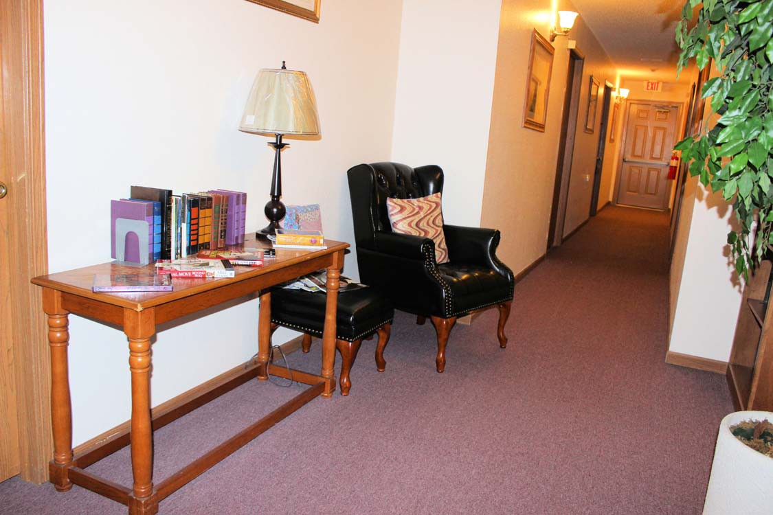 A cozy hallway with a black armchair and a wooden table featuring books and a lamp.