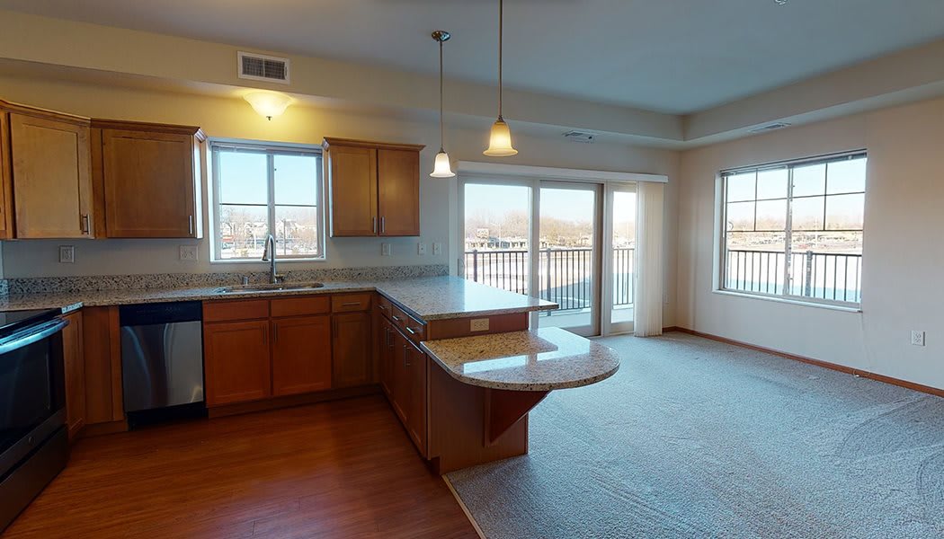 A modern open-plan kitchen with wooden cabinets and a large island countertop, featuring pendant lighting and ample natural light from windows and a sliding door.