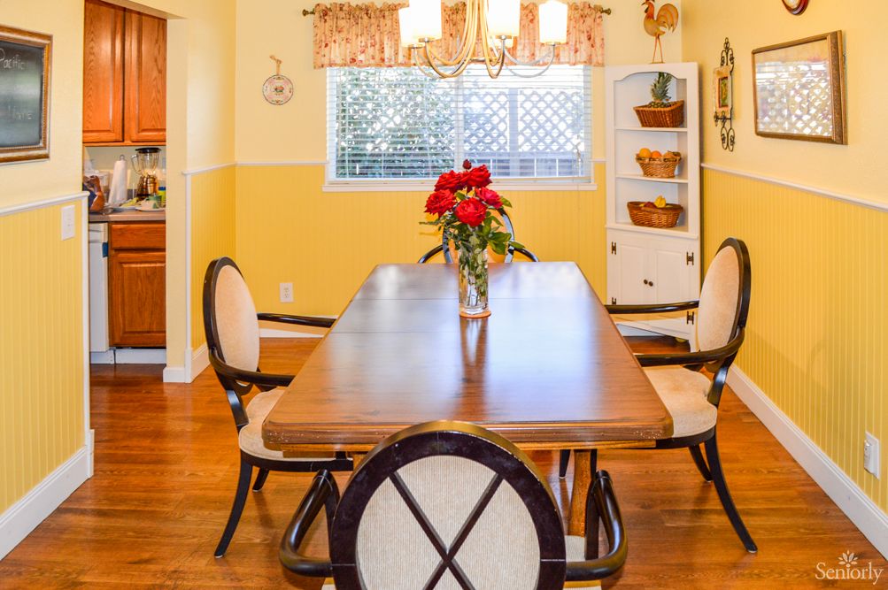 A bright dining room featuring a wooden table set with a vase of red flowers, surrounded by chairs, under a chandelier.