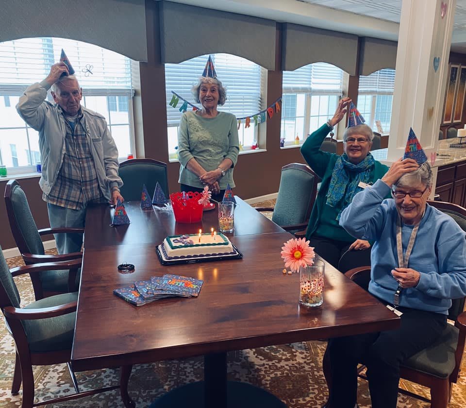 A group of seniors celebrating a birthday indoors, wearing party hats around a table with a cake.