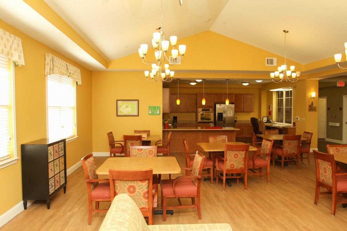 A cozy dining area with warm yellow walls, wooden tables and chairs, and a view into a kitchen.