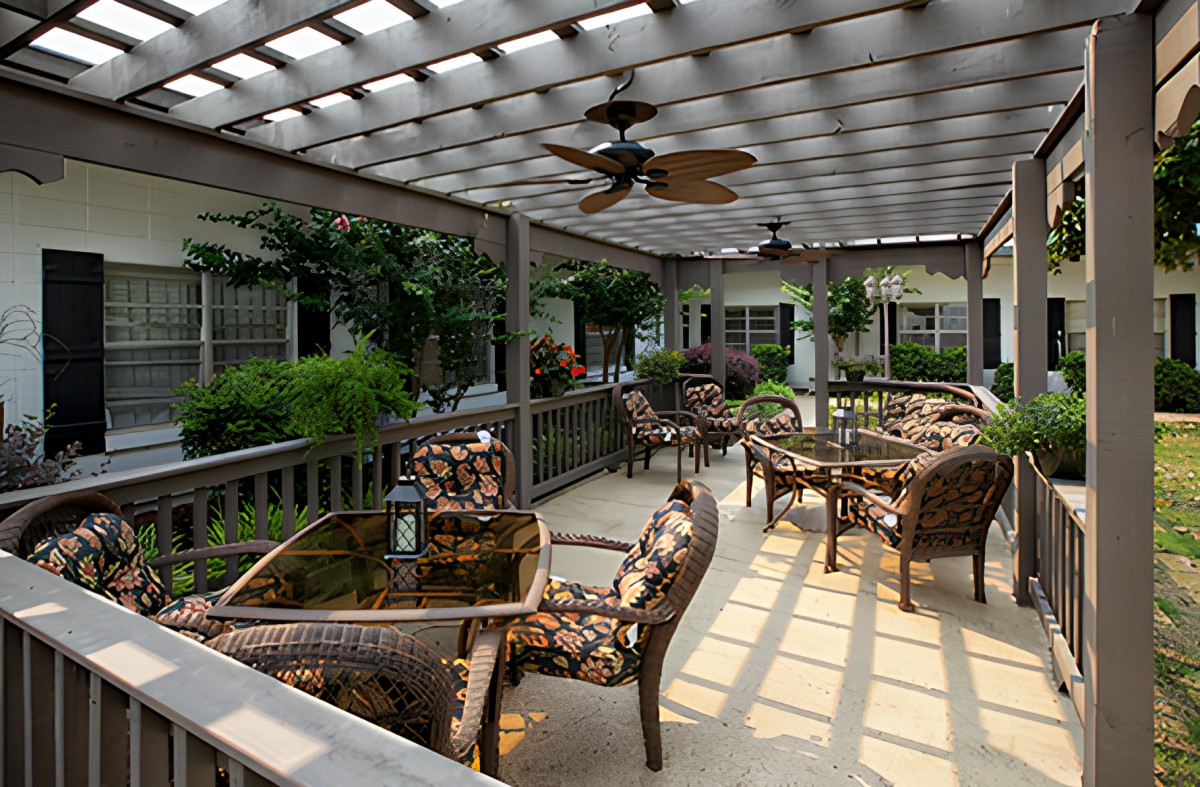 A cozy outdoor patio area featuring wicker furniture under a pergola, with lush greenery and a ceiling fan for comfort.