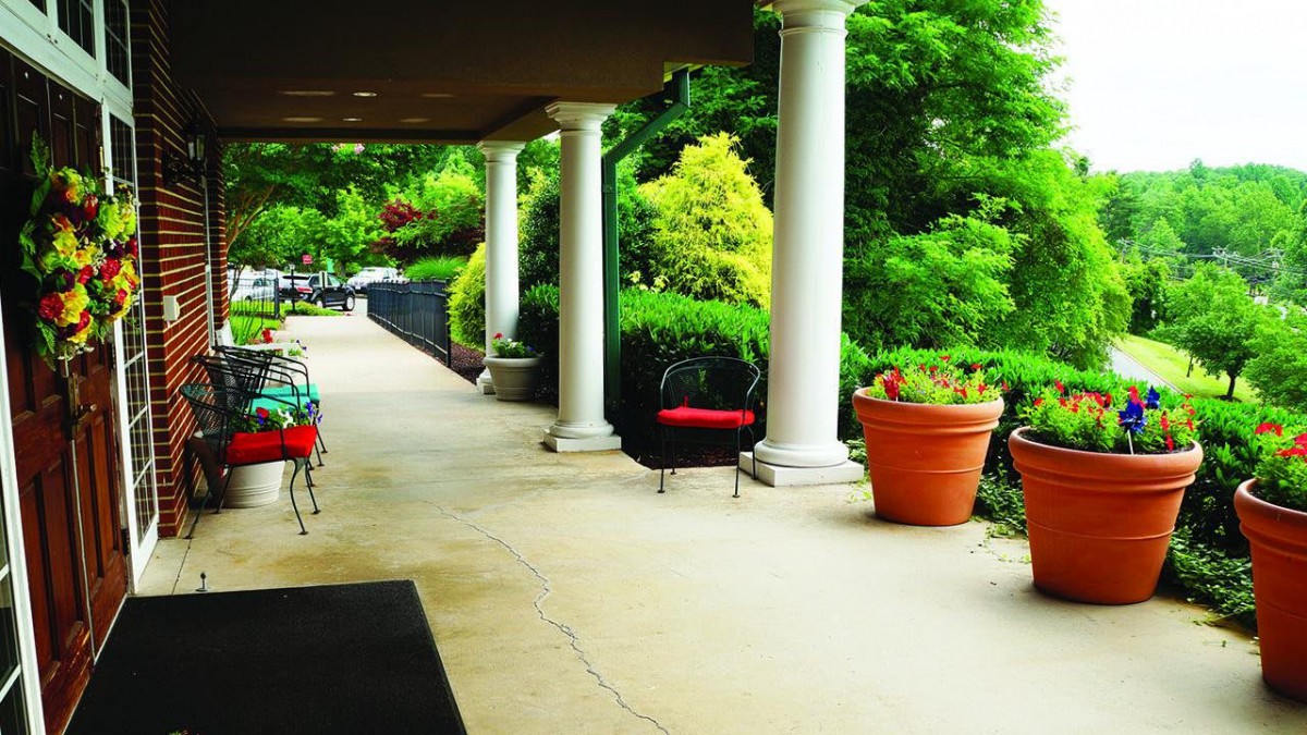 A serene patio with columns and chairs, surrounded by lush greenery and potted plants.