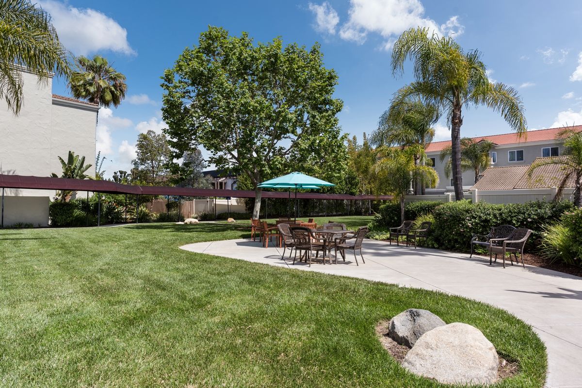 A peaceful residential backyard with a patio set under an umbrella, surrounded by lush lawn and trees.