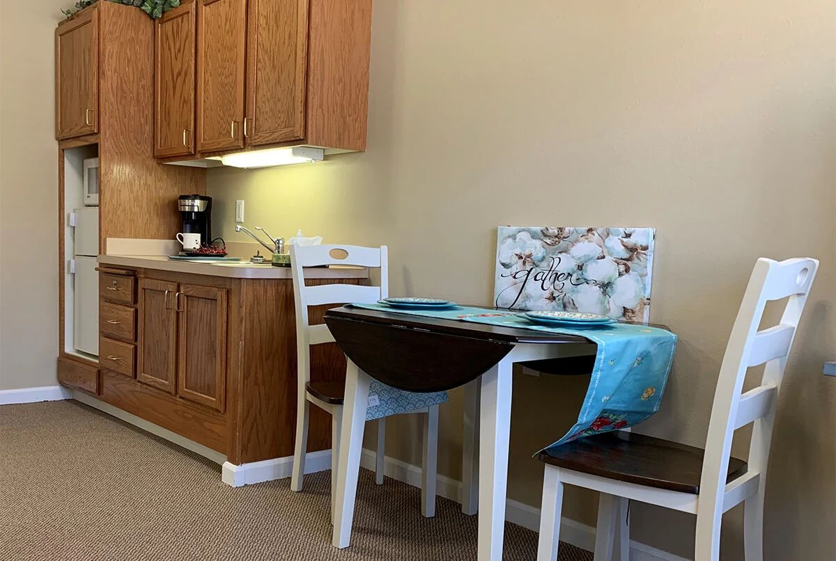 A cozy kitchen nook with a wooden table and chairs, next to a set of cabinets and coffee maker.