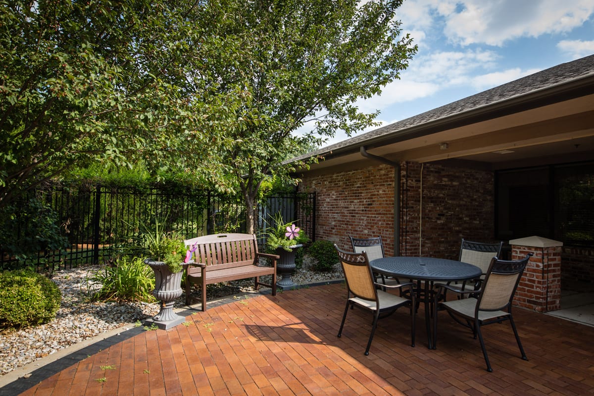 A cozy patio with a bench and table set under a tree, surrounded by brick walls on a sunny day.
