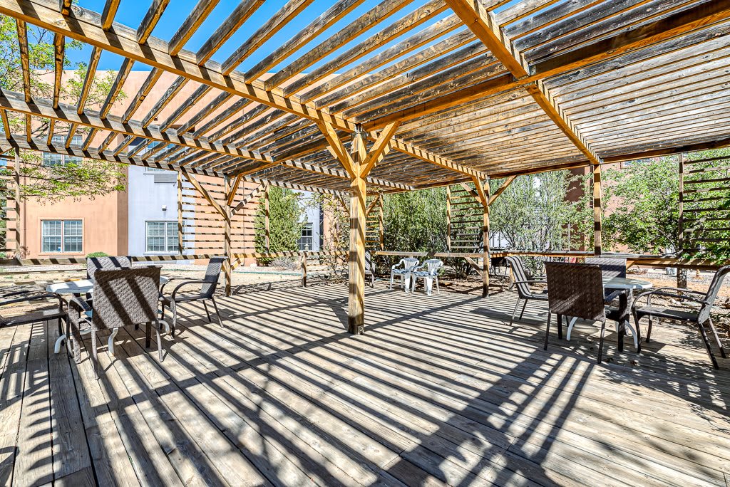 A spacious outdoor patio with a wooden pergola casting intricate shadows, surrounded by seating and greenery.