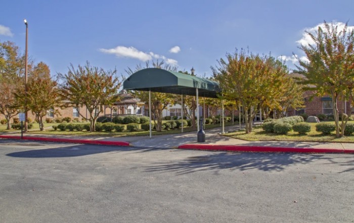 A covered entrance with a green canopy in front of a building surrounded by trees and a parking lot.