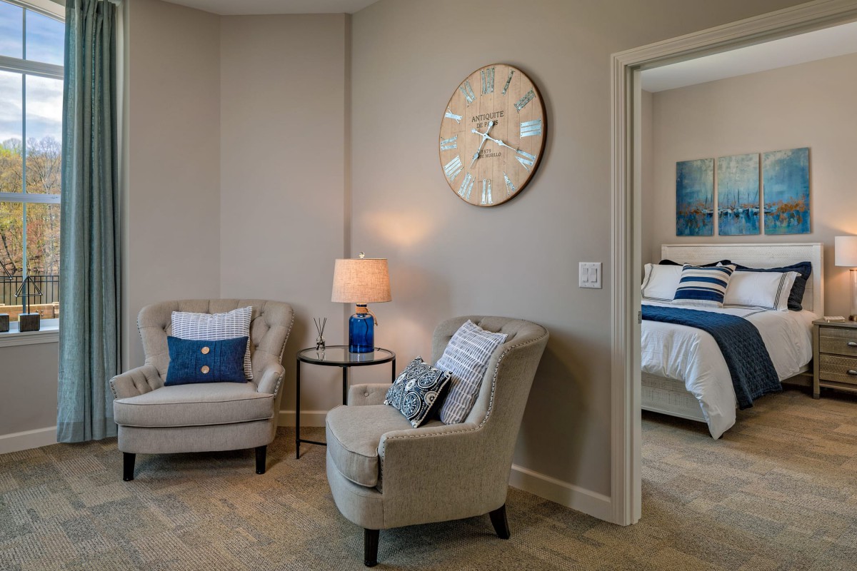 A cozy living room area with elegant armchairs and a stylish lamp, featuring a large wall clock, and an adjacent view into a neatly decorated bedroom.