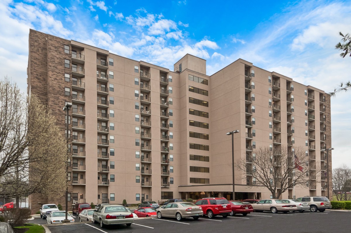 A tall apartment building with a parking lot full of cars under a blue sky.
