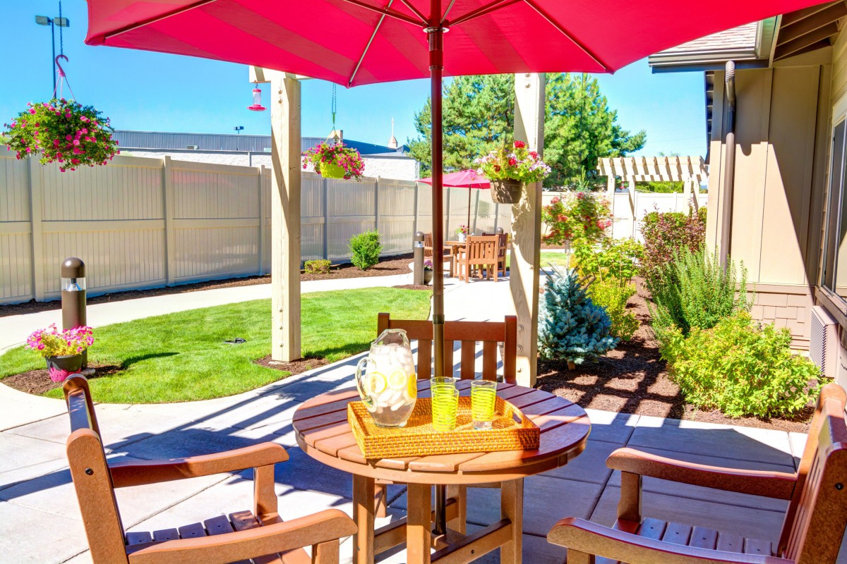 A sunny outdoor patio with a table and chairs under a red umbrella, featuring a pitcher of lemonade.
