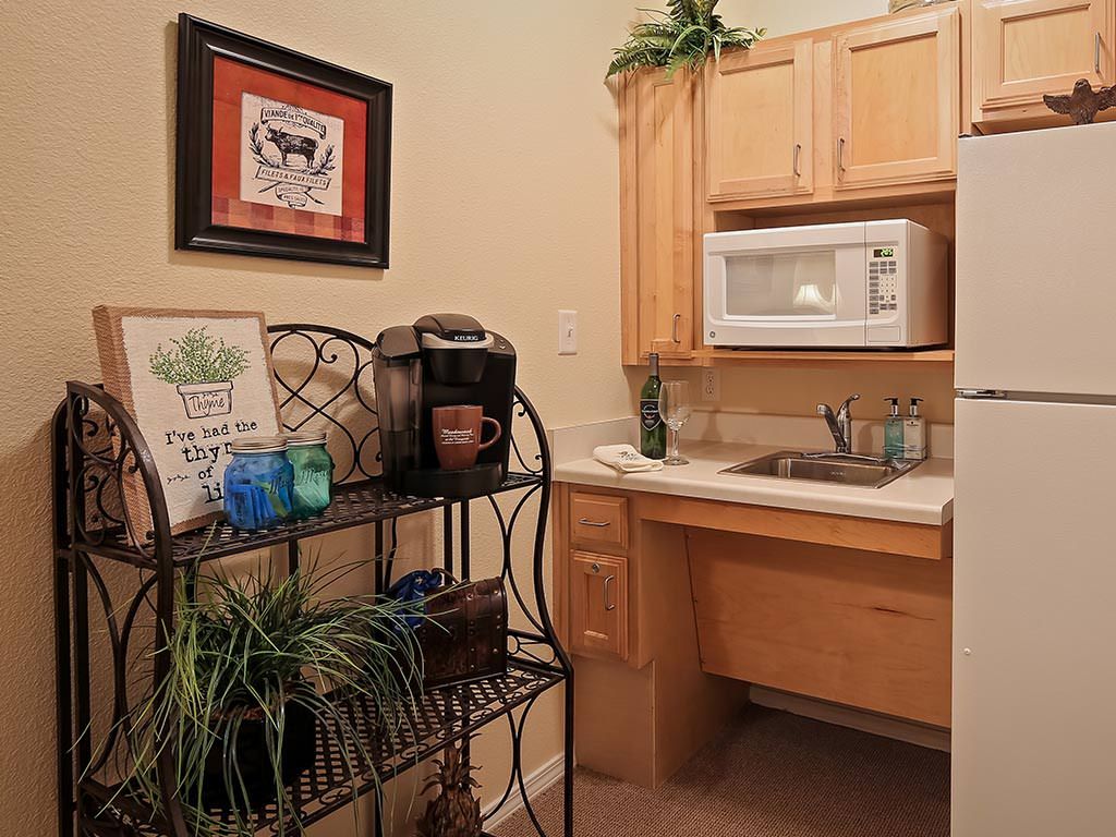 A cozy kitchenette featuring a microwave, sink, wooden cabinets, and a coffee maker on a decorative shelf with plants.