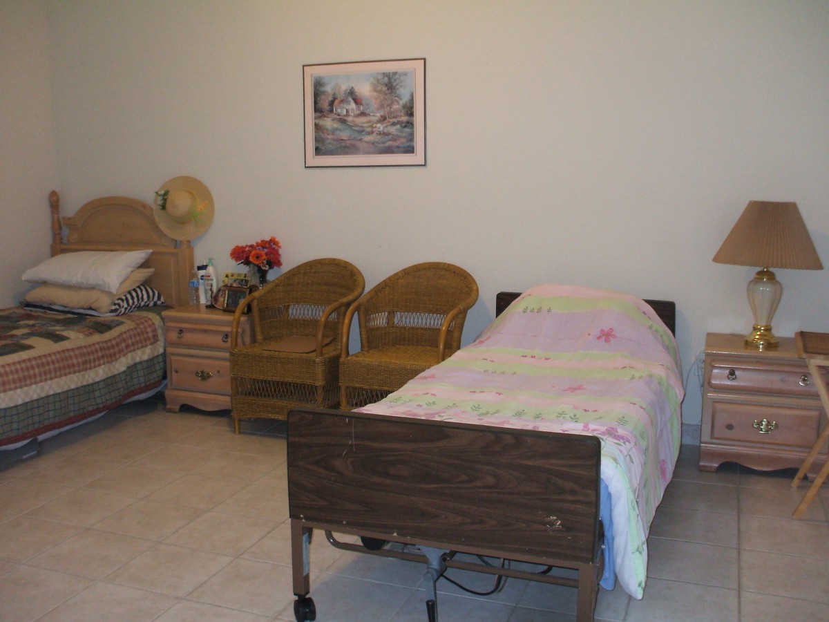 A cozy room featuring two hospital beds, wicker chairs, and a painting on the wall.