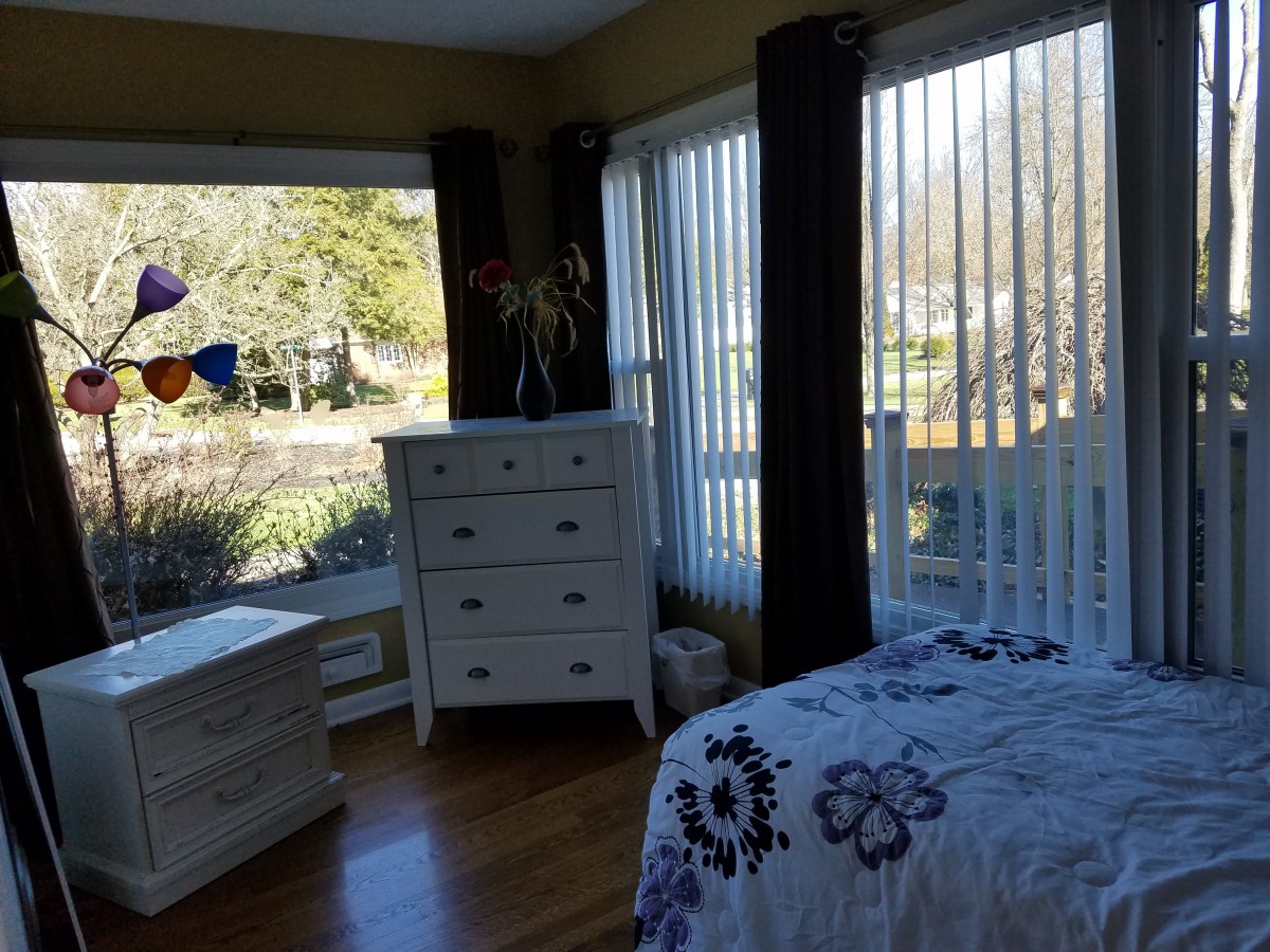 A cozy bedroom with large windows, a white dresser, and a bed decorated with purple floral bedding.